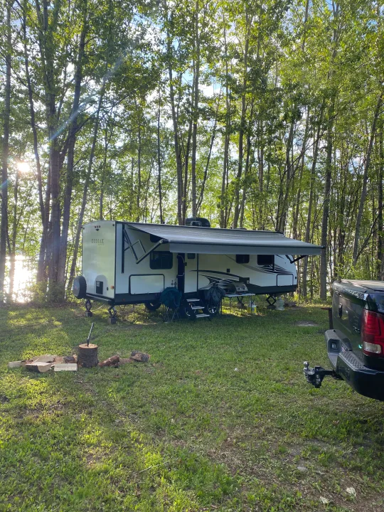 Lakeside RV rental camping with a white travel trailer, campfire, and truck in a sunny wooded area by the water.. Dutchmen Kodiak Ultra Lite 2020