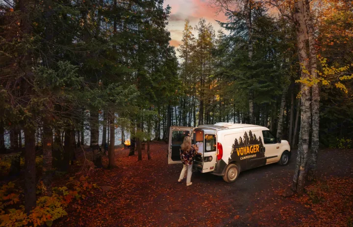 Autumn lakeside camping with an RV rental. Voyager Campervan parked in a fall forest by water, person at back, distant campfire.. Dodge Promaster City 2020