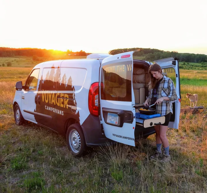 A woman prepares a meal at sunset from a white campervan (RV rental) in a grassy field, with a dog nearby.. Dodge Promaster City 2020