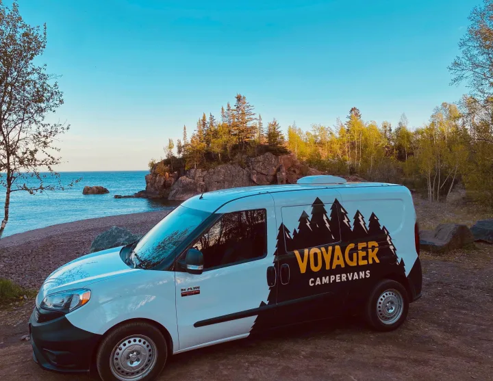 A Voyager campervan RV rental is parked on a rocky shore by a lake, with cliffs and trees in the background under a blue sky.. Dodge Promaster City 2020