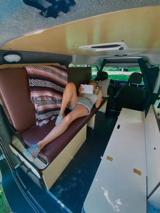 Cozy RV rental interior: A person relaxes, reading a book on a built-in brown bench in this compact camper van.. Dodge Promaster City 2020