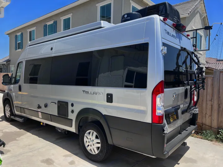 A silver Ram ProMaster Tellaro RV rental, fully equipped with a bike rack and rooftop amenities, parked in a driveway.. Thor Tellaro 2021