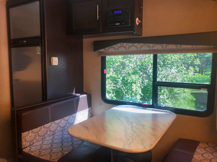Cozy RV rental interior shows a dinette by a large window with green trees outside, plus a fridge and entertainment system.. Dutchmen Kodiak Express 2017