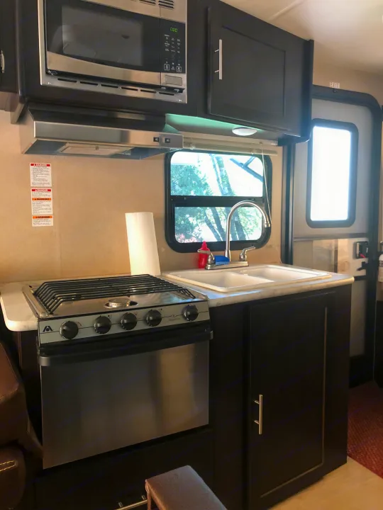 RV rental kitchen: stainless microwave, range/oven, sink, and dark cabinets. Window view & exit door visible.. Dutchmen Kodiak Express 2017