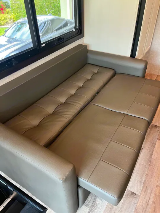 Spacious RV rental interior with a brown convertible sleeper sofa extended for use. A car is visible through the window.. Entegra Vision 2023