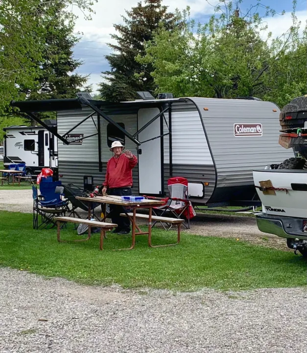 Man waving at his Coleman RV rental in a wooded campground. Picnic table, fishing gear, and truck nearby.. Dutchmen Coleman Lantern LT 2023