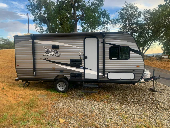 Jayco Jay Flight travel trailer rv rental parked on gravel and dry grass outdoors, with trees in background.. Jayco Jay Flight SLX 2020