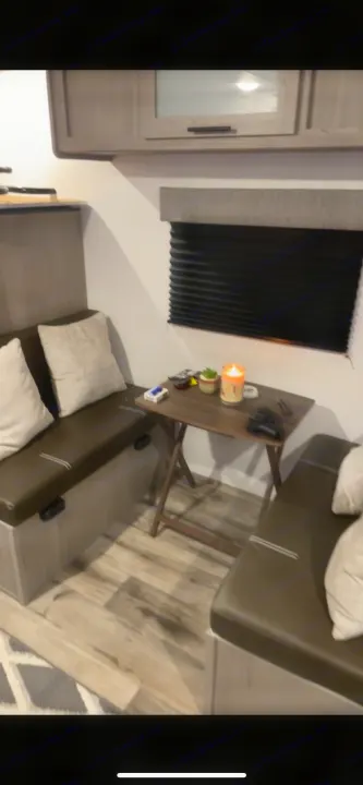 Comfy rv rental interior: A dinette with dark green bench seats, light pillows, a folding wooden table with a lit candle, and window blinds.. Coleman Lantern 17B 2023