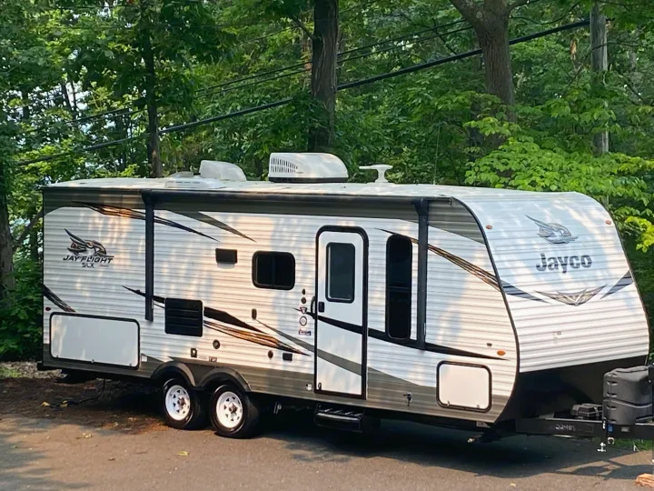 A white and gray Jayco travel trailer (RV) with striped graphics is parked among trees. Ideal for your next RV rental adventure.. Jayco Jay Flight SLX 2019