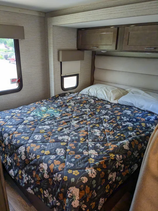 Cozy RV rental bedroom features a dark floral comforter, padded headboard, pillows, windows, and overhead cabinets.. Winnebago Minnie Winnie 2022