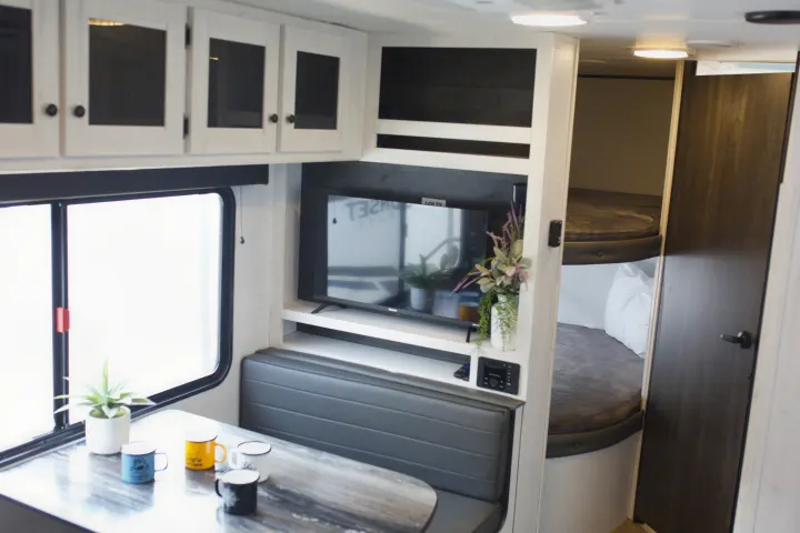 Modern rv rental interior: Light wood dinette, TV, overhead cabinets. Cozy bunk beds are visible in the background.. Heartland Trail Runner 2022