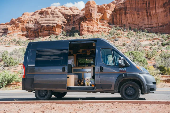 Pet-friendly rv rental van: a gray camper with its side door open, revealing two dogs & custom interior, parked near red rocks.. Ram Promaster 2022