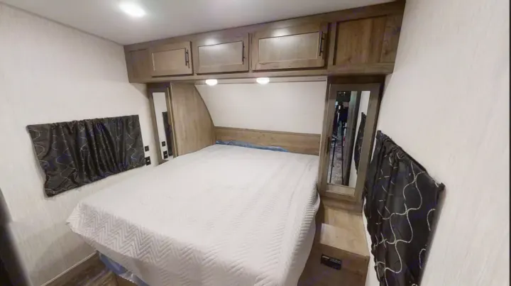 Cozy RV rental bedroom featuring a bed with a white comforter, wooden cabinets, wall lights, and dark privacy curtains.. Forest River Cherokee Wolf Pack 2020