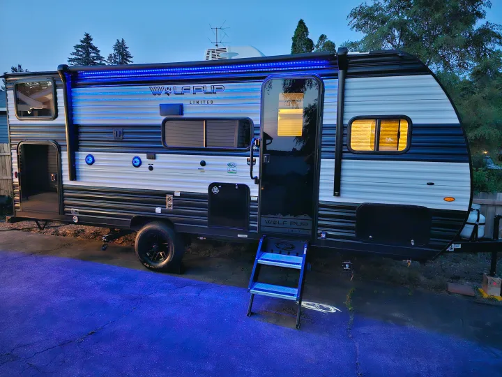 This illuminated RV rental, a Wolf Pup travel trailer, features striking blue LED lighting on its awning and steps at dusk.. Forest River Wolf pup 17JW 2023