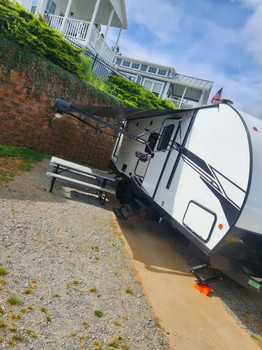 White RV rental with awning extended, parked on gravel next to a multi-story house flying an American flag.. Forest River Tracer 2022