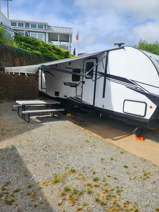 An RV rental, a white travel trailer, parked at a gravel campsite with a picnic table and extended awning. Perfect for camping.. Forest River Tracer 2022