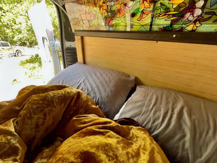 Cozy RV rental bed with gold blanket, gray pillows, stained glass & open door view.. Dodge Ram ProMaster 2500 High Top 2018