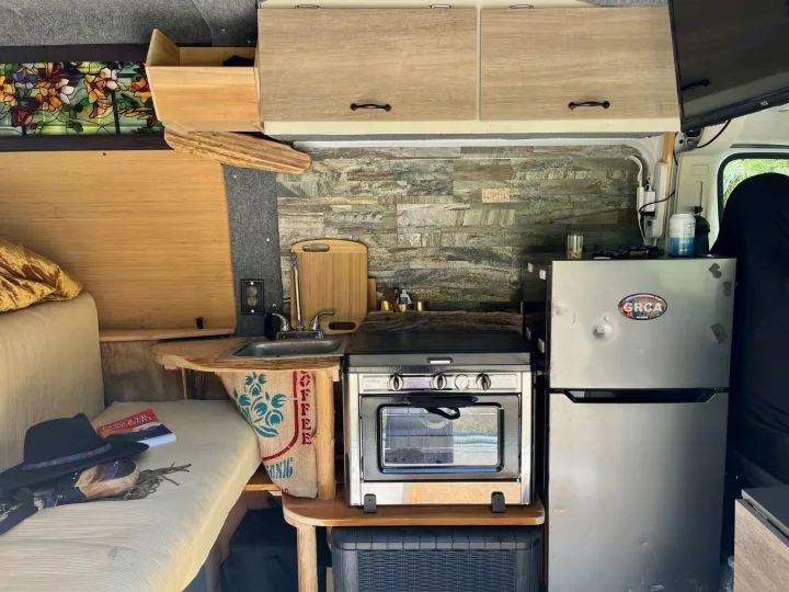 RV rental interior: compact kitchen with sink, stove, oven, fridge. Features seating, custom stained glass. Perfect mobile setup.. Dodge Ram ProMaster 2500 High Top 2018