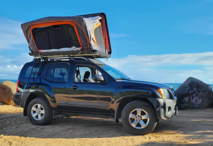 Dark SUV with rooftop tent parked on a sandy beach. Ready for your RV rental adventure by the ocean.. Nissan Exterra 2011