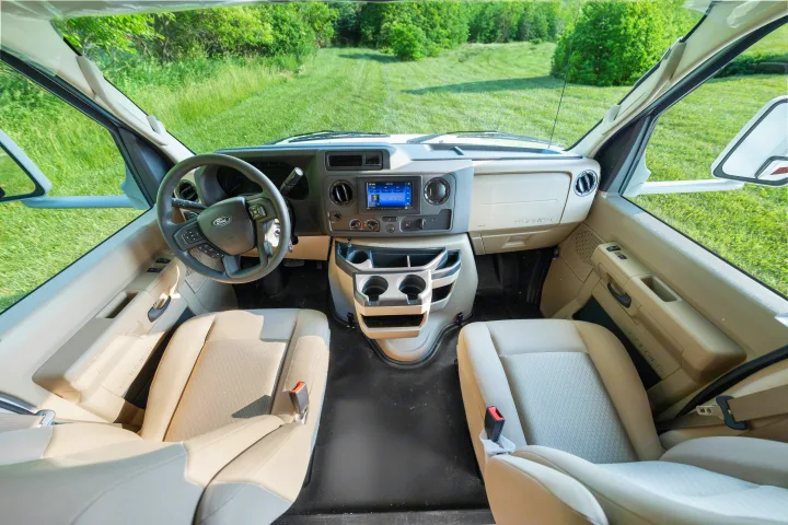 This RV rental features a comfortable, modern cockpit with a Ford dashboard and clear windshield view of a grassy field.. Thor Coleman 2023