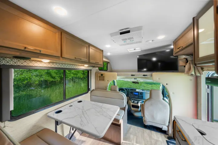 Comfortable RV rental interior with a dining area, overhead cabinets, and a large window showcasing green scenery.. Thor Coleman 2023