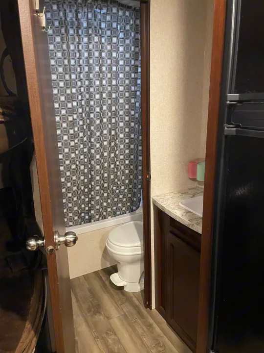 Inside an RV rental: a compact bathroom with shower, toilet, and sink, visible through an open door, beside a refrigerator.. Jayco Jay Flight SLX 2019