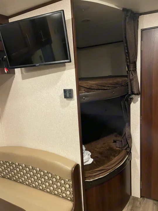 RV rental interior with a TV above a sofa, next to bunk beds with a privacy curtain.. Jayco Jay Flight SLX 2019