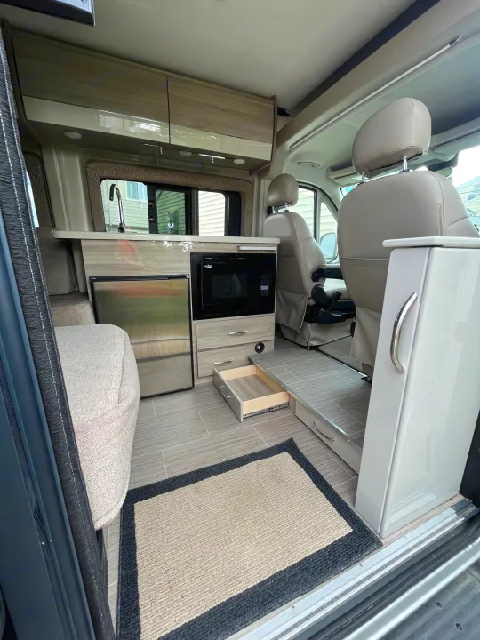 Modern rv rental interior: kitchen with fridge, microwave, seating, and swivel chairs. Storage drawer open on grey tiled floor.. Thor Sequence 2020