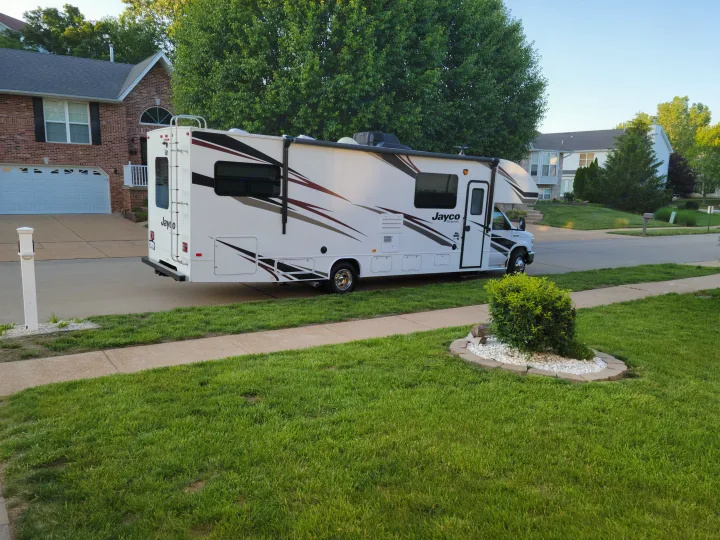 A spacious white Jayco RV rental sits parked curbside on a quiet, tree-lined residential street next to a house.. Jayco Redhawk 2020