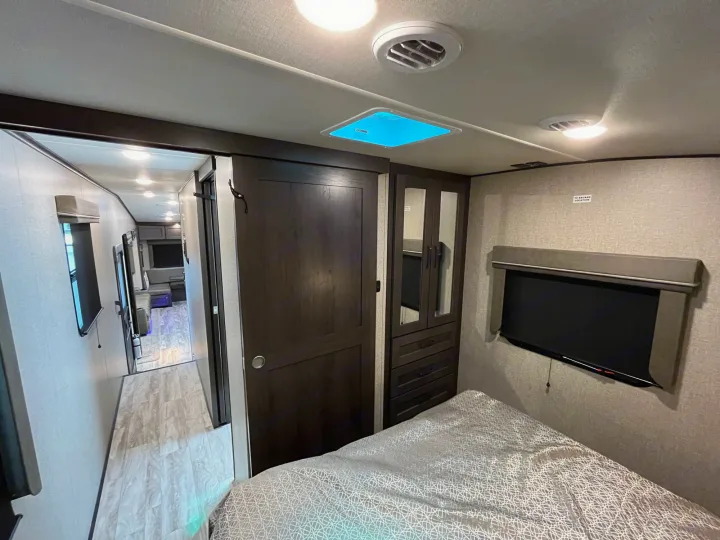 Inside an RV rental, a cozy bedroom with storage and TV connects to a bright hallway, showcasing compact living comfort.. Grand Design Reflection 2022