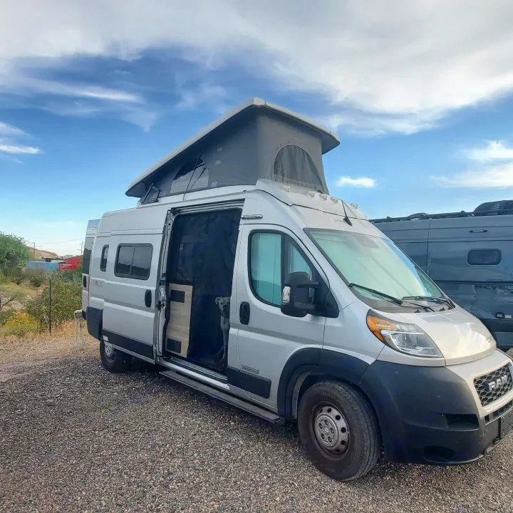 Silver camper van for RV rental, with pop-top roof up & side door open revealing a dog inside, parked on gravel.. Winnebago Solis 2022
