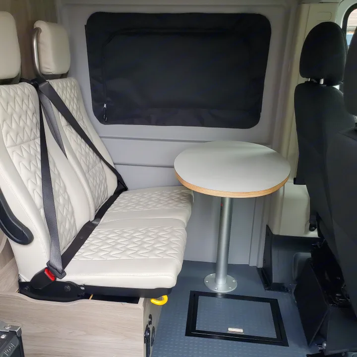 RV rental interior featuring light patterned bench seats with seatbelts, a round table, and dark window blind.. Winnebago Solis 2022