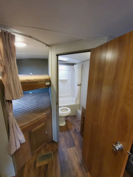 Interior of an RV rental: showing two bunk beds with curtains, and a compact bathroom with a toilet and shower.. Keystone 27BHS 2017