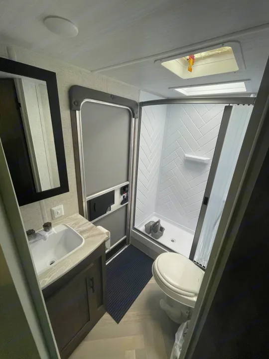 Inside an RV rental bathroom: a modern vanity, mirror, toilet, and a compact shower with white herringbone tiles.. Forest River Wildwood X-Lite 2021