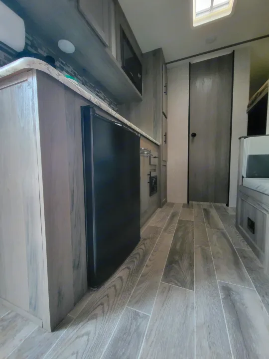 Interior view of an RV rental kitchen and living space, featuring wood-look flooring, a compact fridge, and overhead cabinets.. Gulf Stream Ameri-Lite Super Lite 2021
