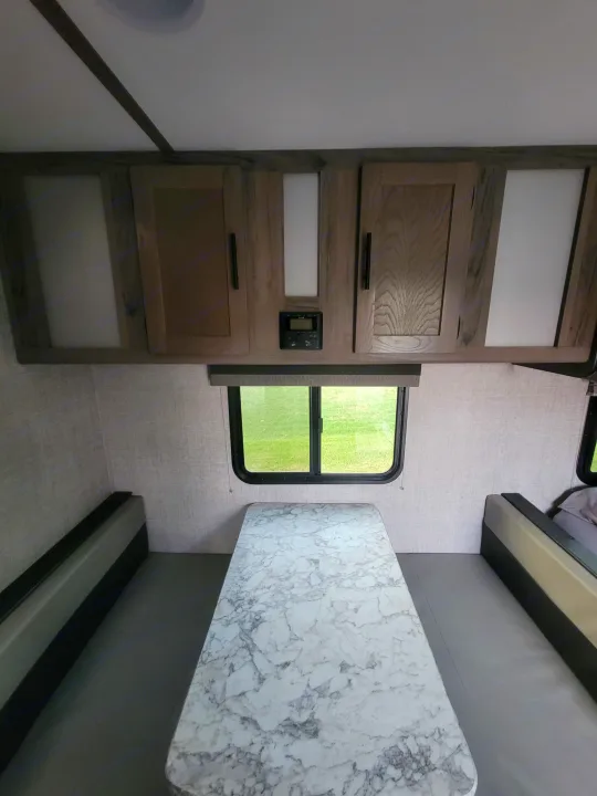 Inside an RV rental, a dinette features a marble-pattern table, gray benches, overhead cabinets, and a window view.. Gulf Stream Ameri-Lite Super Lite 2021