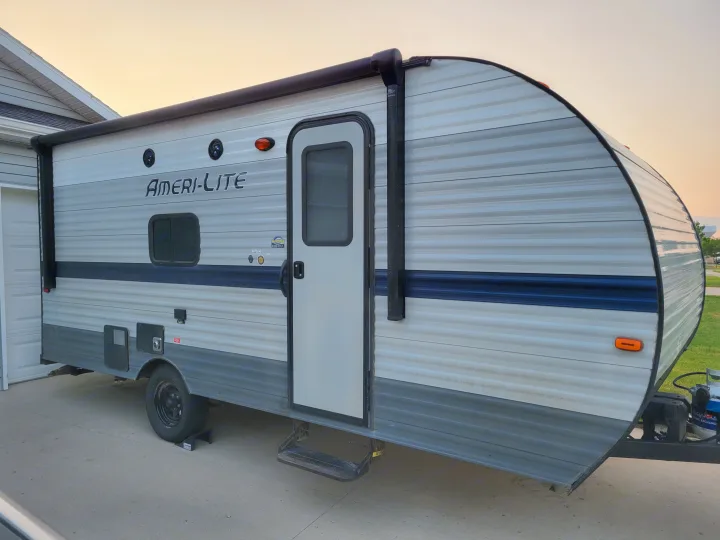 A light gray and blue travel trailer, an RV rental, is parked on a concrete driveway, ready for your next adventure.. Gulf Stream Ameri-Lite Super Lite 2021