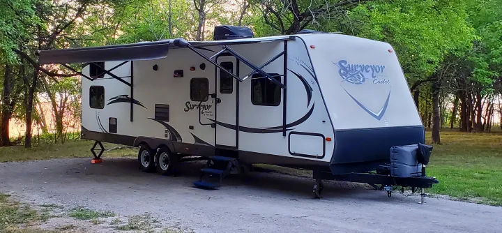 An RV rental, a Surveyor Cadet travel trailer, parked for camping in a wooded outdoor setting with awning extended.. Forest River Surveyor 2015