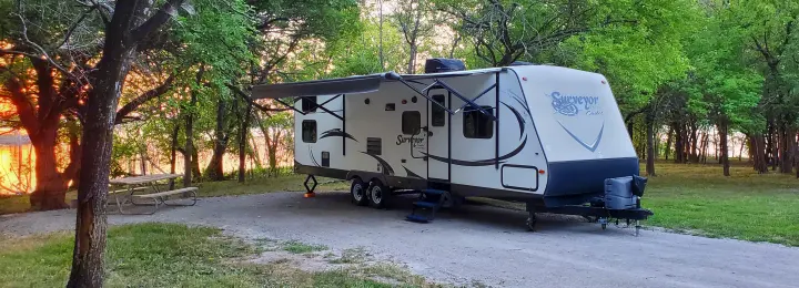 RV rental trailer parked at a serene lakeside campsite with a picnic table, bathed in sunset glow through the trees.. Forest River Surveyor 2015