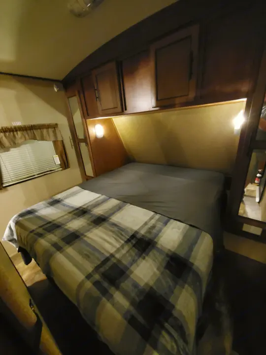 Inside an RV rental, a comfortable bedroom with plaid blanket, overhead cabinets, and a full-length mirror.. Forest River Surveyor 2015