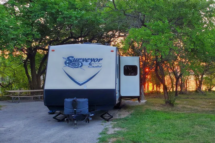 Enjoying an RV rental. A travel trailer parked at a tree-filled campsite with bright orange sunset light peeking through.. Forest River Surveyor 2015