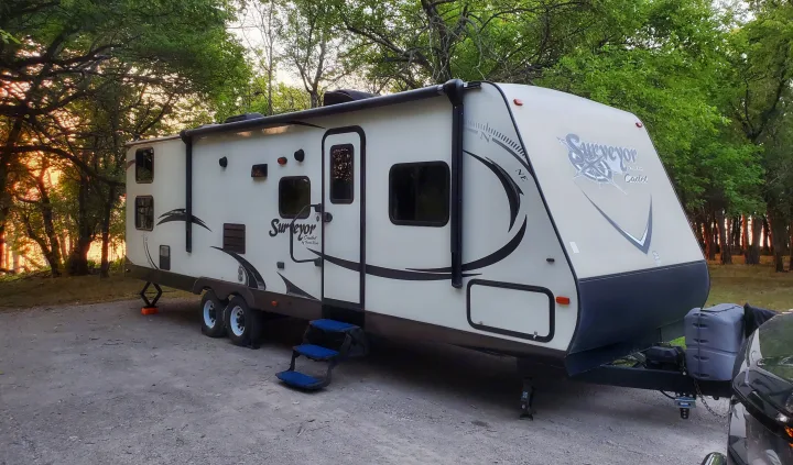 A beige travel trailer RV rental sits on gravel, surrounded by trees at sunset, perfect for camping.. Forest River Surveyor 2015