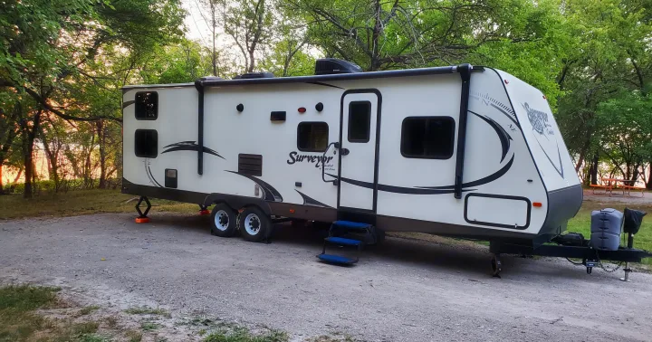 White travel trailer RV rental parked at a wooded campsite during sunset, ready for camping adventures.. Forest River Surveyor 2015