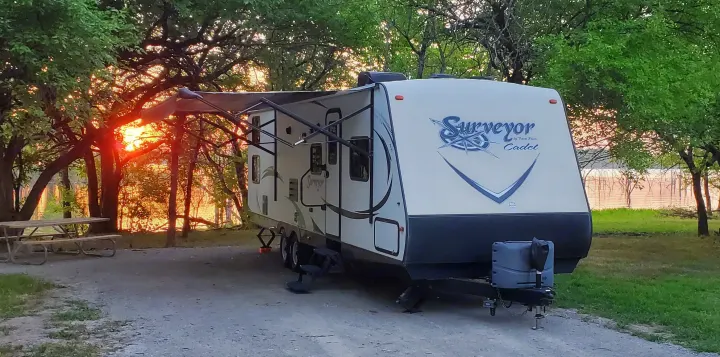 RV rental travel trailer parked at a scenic campsite. Golden sunset over a lake, visible through trees, creates a warm glow.. Forest River Surveyor 2015