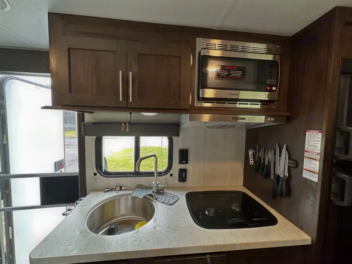 Compact RV rental kitchen features dark wood cabinets, a stainless steel sink, two-burner cooktop, and overhead microwave.. Forest River Cherokee Wolf Pup 2020