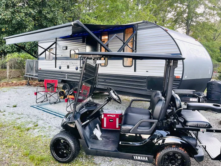 RV rental setup: A modern travel trailer with awning and lights, parked alongside a black golf cart at a gravel campsite.. Forest River Cherokee Grey Wolf 2019