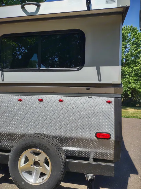 Side and rear of a compact travel trailer with a window, diamond plate panel, and spare tire, ready for an RV rental.. A Liner A Liner Popup Trailer 2022