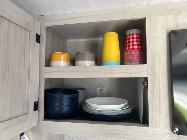RV rental kitchen cabinet reveals neatly stacked plates, bowls, and colorful mugs & cups, essential for travel.. East to West Silver Lake LE 2023