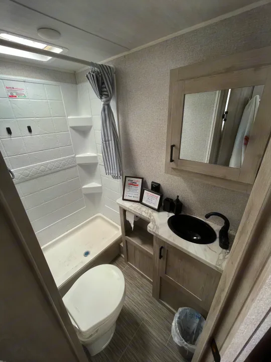 This clean rv rental bathroom features a compact shower, toilet, and a vanity with a black sink and mirror.. East to West Silver Lake LE 2023