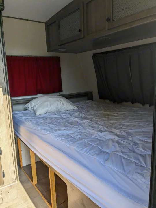 RV rental interior: A bed with white bedding, curtains, and wood-toned cabinets in a cozy camper sleeping area.. Coachmen Apex Nano 2021